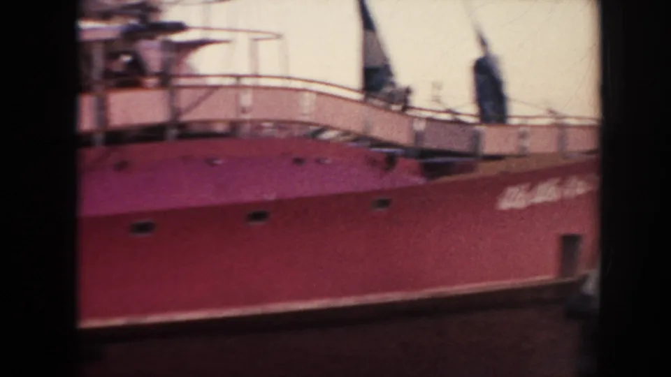 A red-hulled ship is filmed from the dock, with railings, deck structures, and soft 8mm motion blur visible.