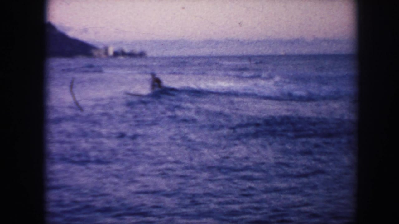 1961 Hawaii ocean view with a distant surfer in the water, low waves, and a shoreline or headland at left.