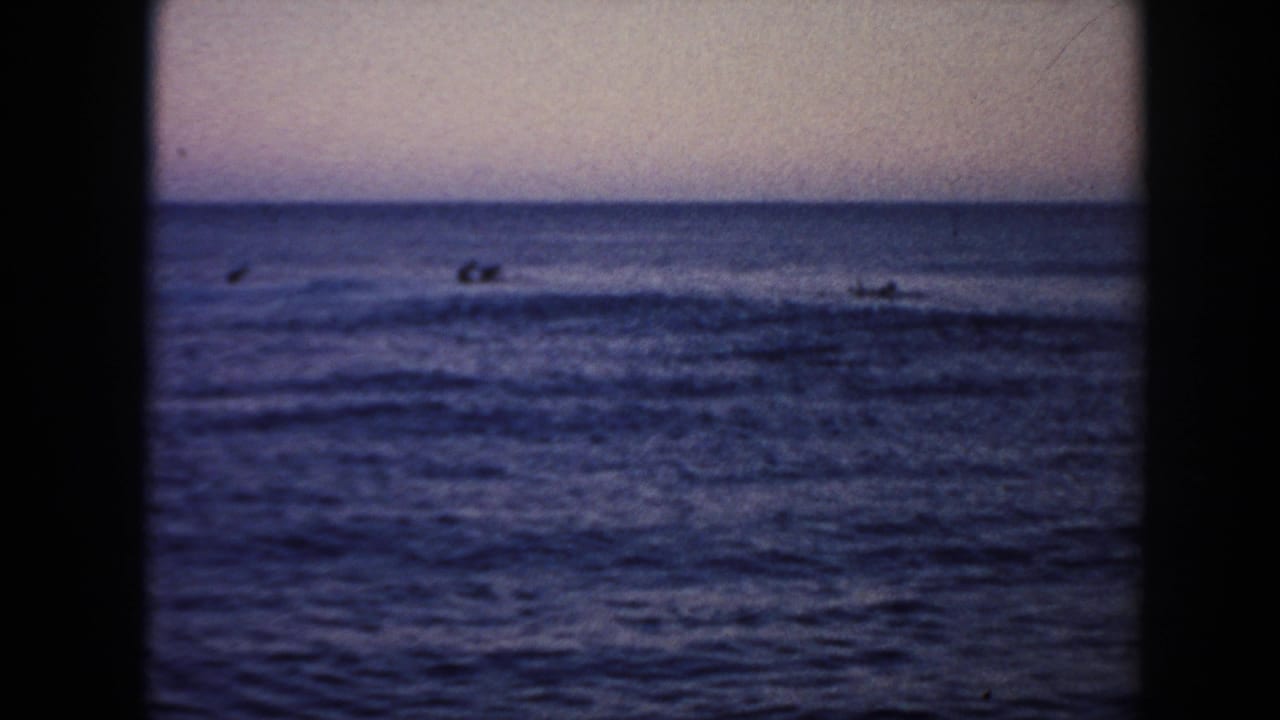 1961 Hawaii 8mm frame with several distant surfers or swimmers beyond low breaking waves under a muted sky.
