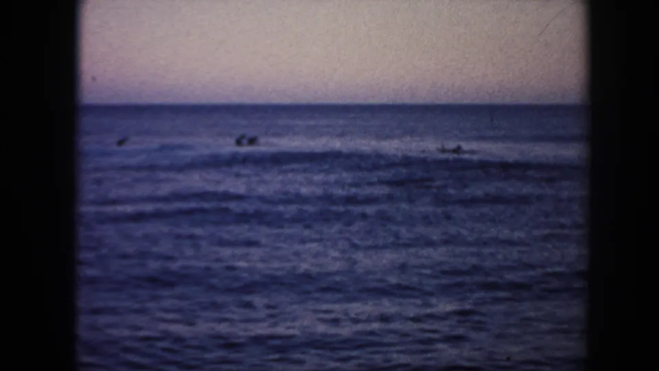 1961 Hawaii 8mm frame with several distant surfers or swimmers beyond low breaking waves under a muted sky.