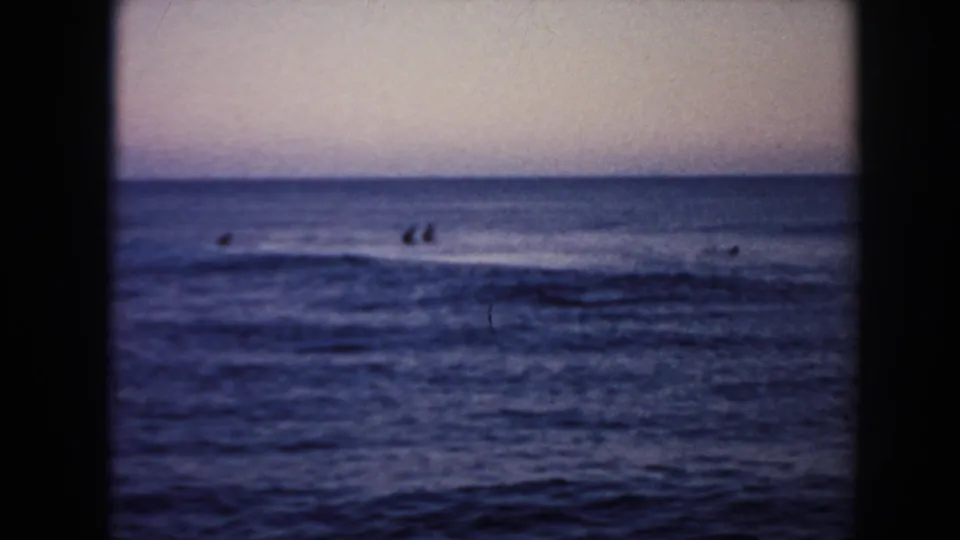 1961 Hawaii 8mm frame showing small surfer silhouettes offshore beneath a muted purple-pink sky and dark ocean surface.