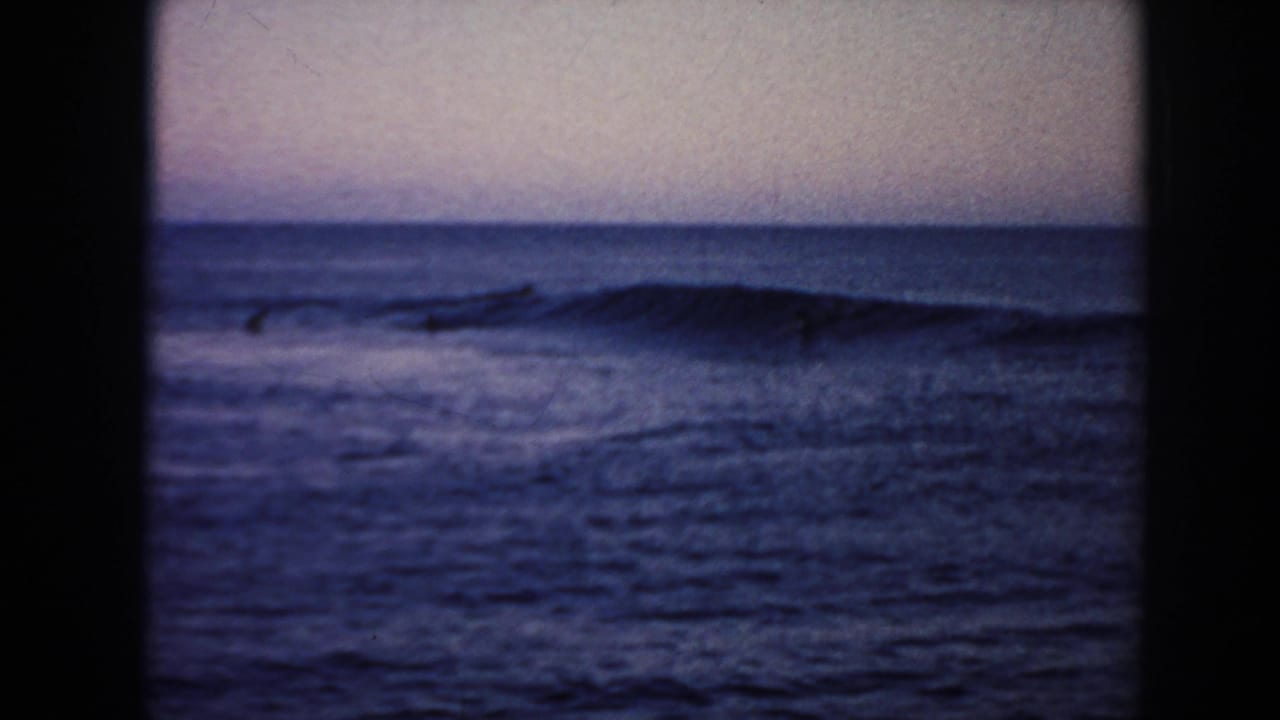 1961 Hawaii 8mm frame with a low wave forming across the right side and small surfer silhouettes scattered offshore.