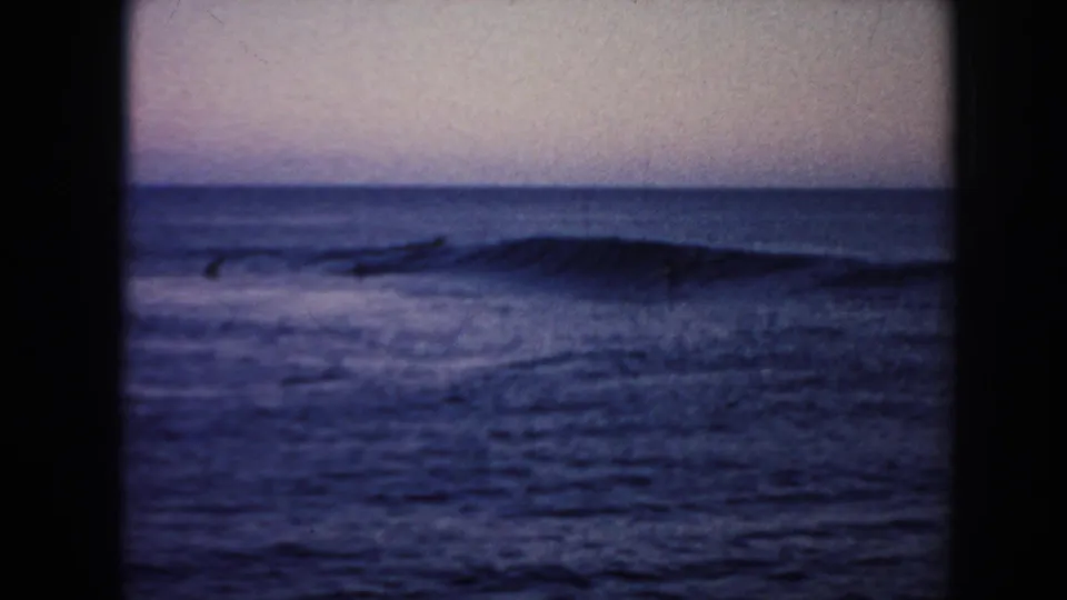 1961 Hawaii 8mm frame with a low wave forming across the right side and small surfer silhouettes scattered offshore.