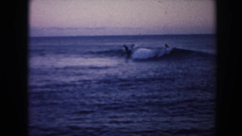 1961 Hawaii 8mm frame showing silhouetted surfers near a white cresting wave, filmed from shore in fading light.
