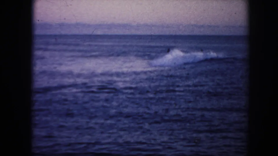 1961 Hawaii 8mm frame of a modest wave breaking at dusk, with surfers appearing as small dark shapes near the whitewater.