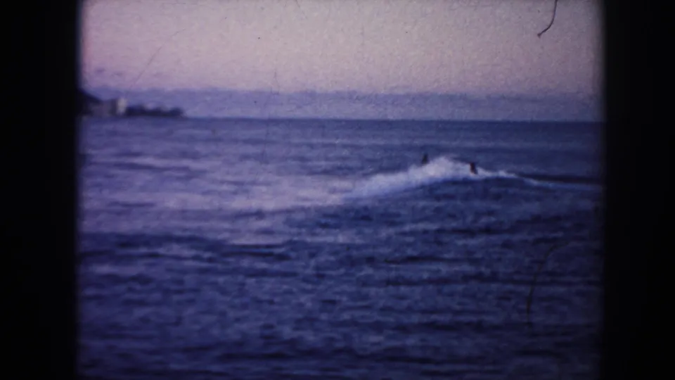 1961 Hawaii 8mm frame showing a small surfer or swimmer in blue ocean waves with a low distant shoreline under a dim sky.