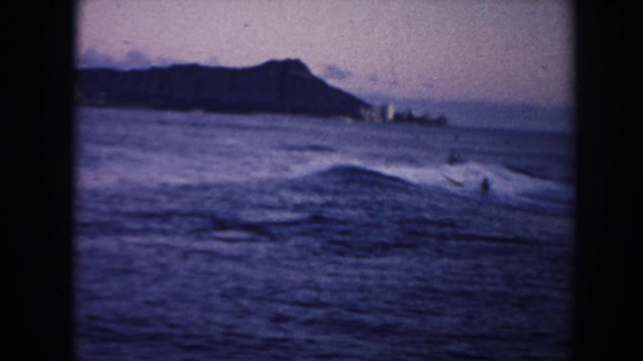 1961 Hawaii 8mm frame with Diamond Head in the background, low surf in the foreground, and distant surfers or swimmers in the water.