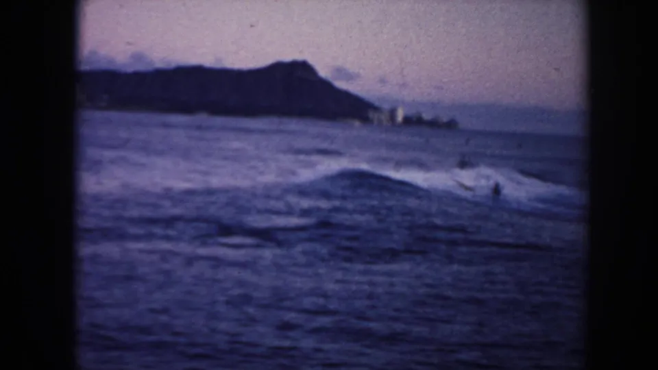 1961 Hawaii 8mm frame with Diamond Head in the background, low surf in the foreground, and distant surfers or swimmers in the water.