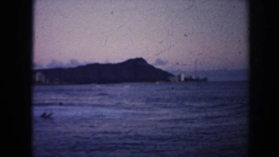 1961 Hawaii 8mm frame showing Diamond Head, distant shoreline buildings, and small surfers or swimmers in the foreground water.