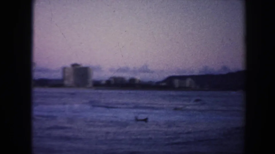 1961 Hawaii 8mm frame looking across the water toward distant buildings and low hills, with a small dark boat-like or surfer shape near the foreground.