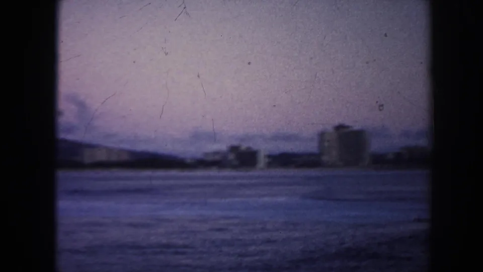 1961 Hawaii home-movie frame looking across calm water toward a distant shoreline, low buildings, and a dark mountain ridge.