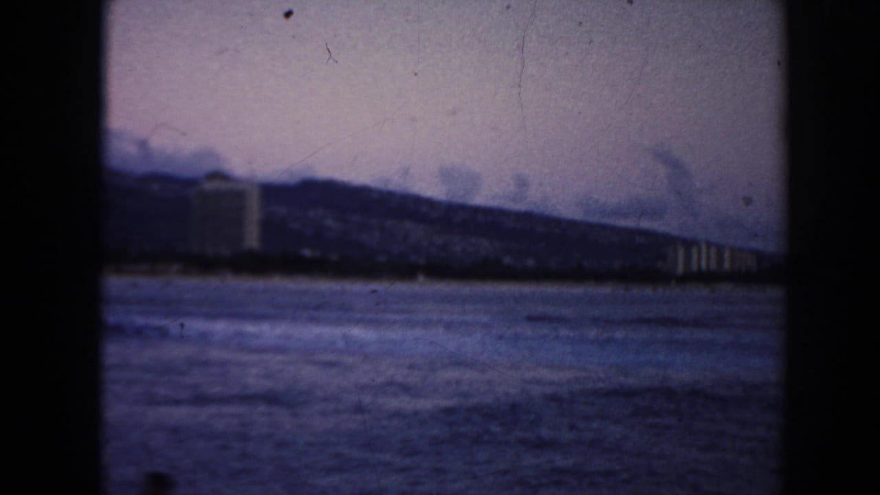 1961 Hawaii coastal frame with open water in the foreground, distant buildings, and a ridge line under a muted sky.