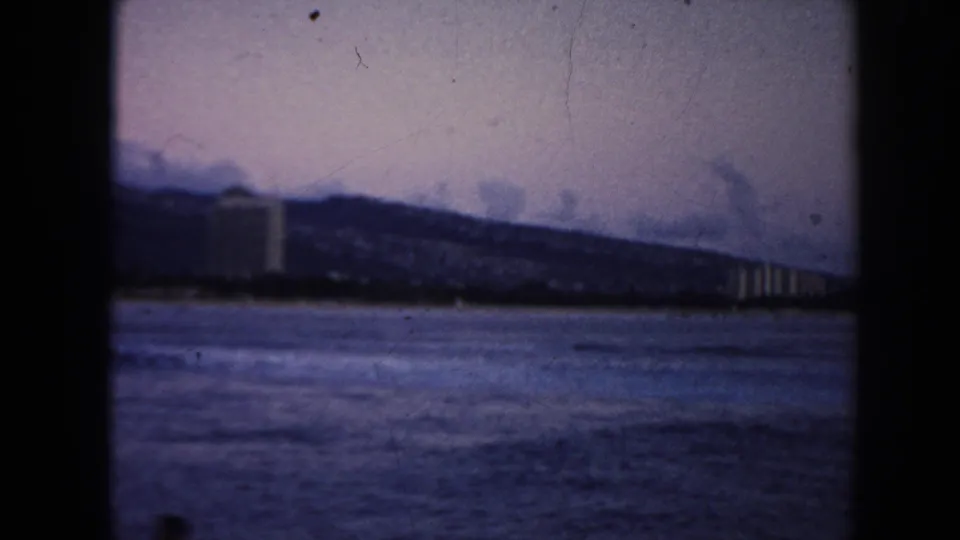 1961 Hawaii coastal frame with open water in the foreground, distant buildings, and a ridge line under a muted sky.