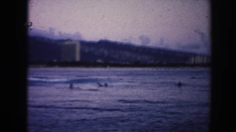 1961 Hawaii 8mm frame showing a waterfront, distant buildings, mountain slope, and small birds or people on the water.