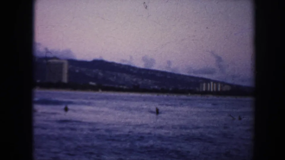 1961 Hawaii home-movie frame of coastal water, a distant ridge, shoreline development, and small indistinct figures on the water.