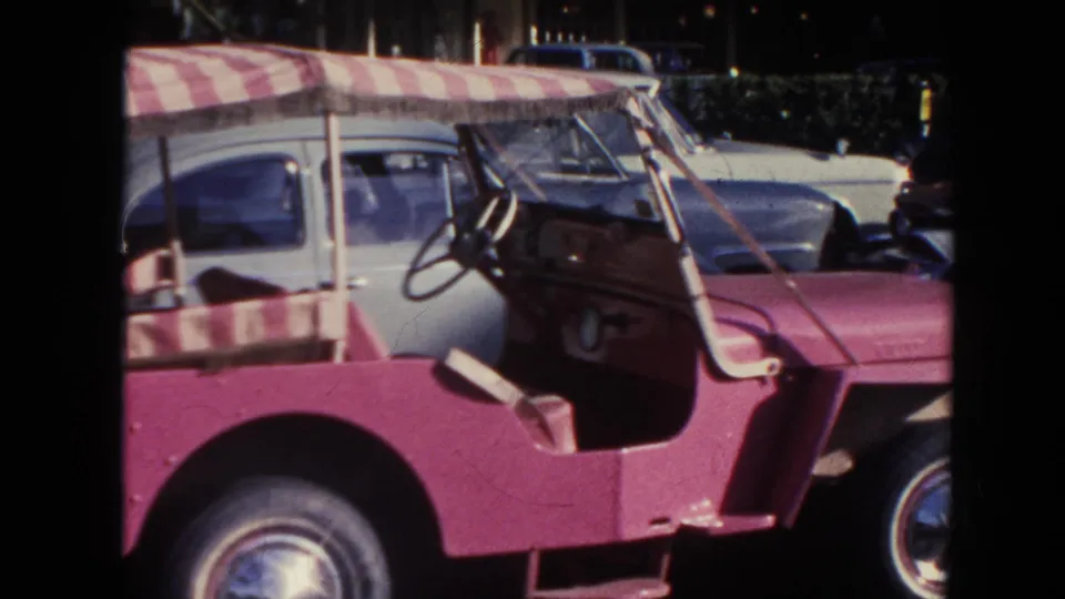 1961 Hawaii frame showing the open pink jeep interior, steering wheel, striped seat, and adjacent light blue car.