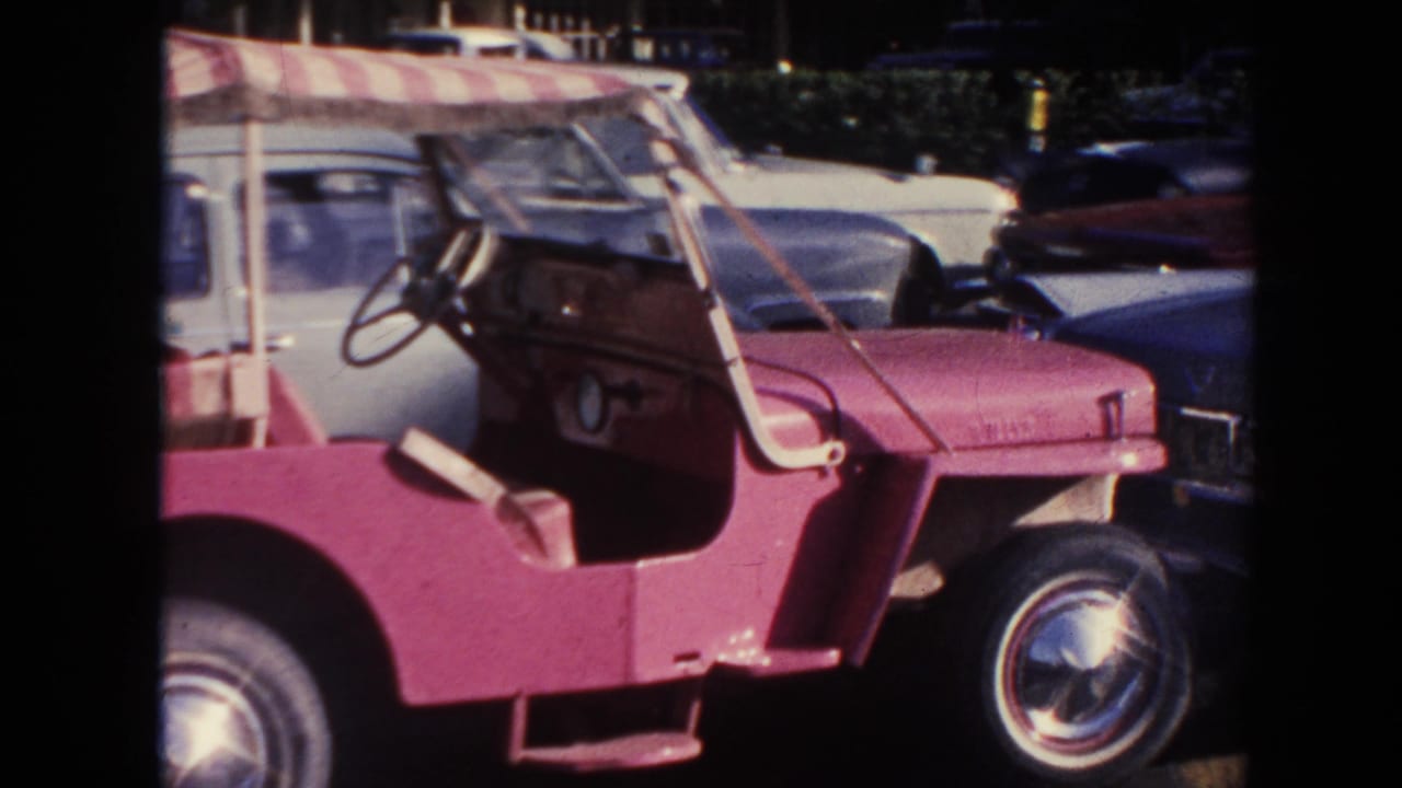 1961 Hawaii 8mm frame showing a pink open buggy with a striped canopy and nearby period cars in a parking area.