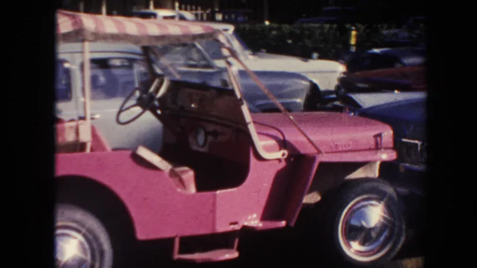 1961 Hawaii 8mm frame showing a pink open buggy with a striped canopy and nearby period cars in a parking area.