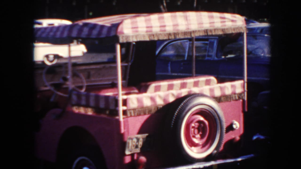 1961 Hawaii home-movie frame showing a pink open vehicle with a red-and-white canopy and spare tire near parked period cars.