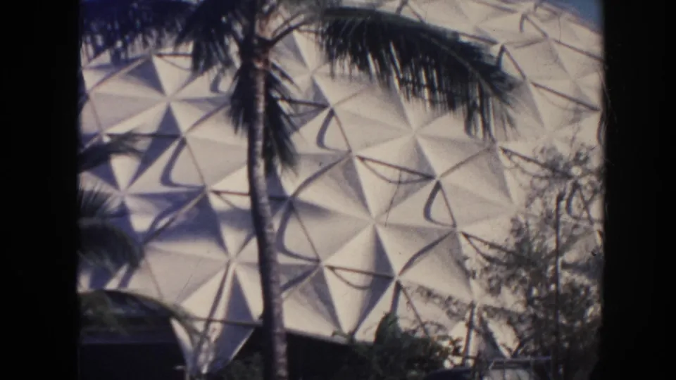 1961 Hawaii home-movie view of a palm trunk crossing a white geometric dome facade with parked cars below.