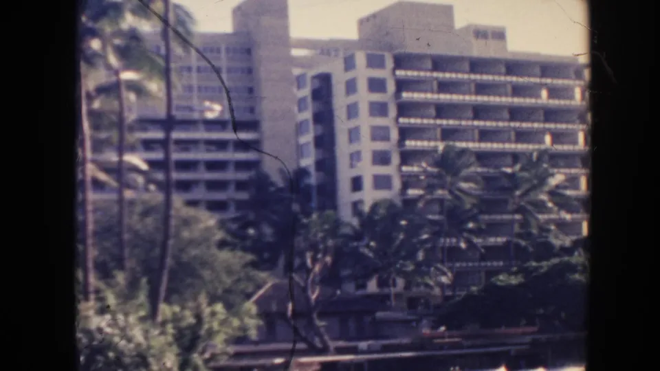 1961 Hawaii resort exterior with a multi-story hotel building, rows of balconies, palm trees, and soft 8mm color.