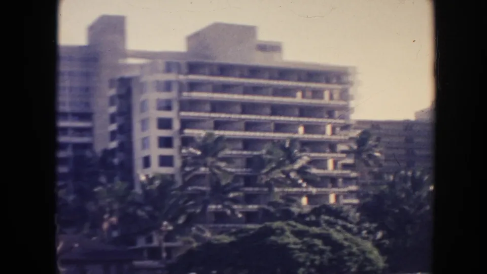1961 Hawaii home-movie frame showing modern multi-story buildings partly screened by palm trees and foliage.