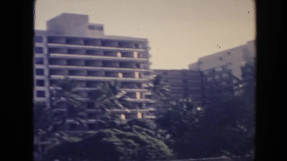 1961 Hawaii frame showing a multi-story building with balcony rows behind palms and dense roadside planting.