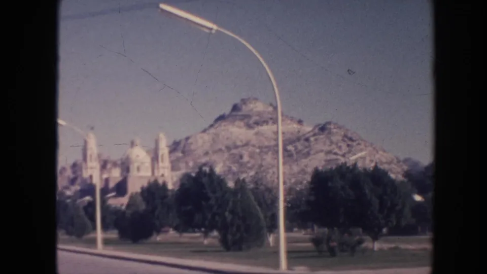 1961 Hawaii 8mm frame showing a rocky mountain behind a distant church-like building, trees, grass, road, and curved streetlight.