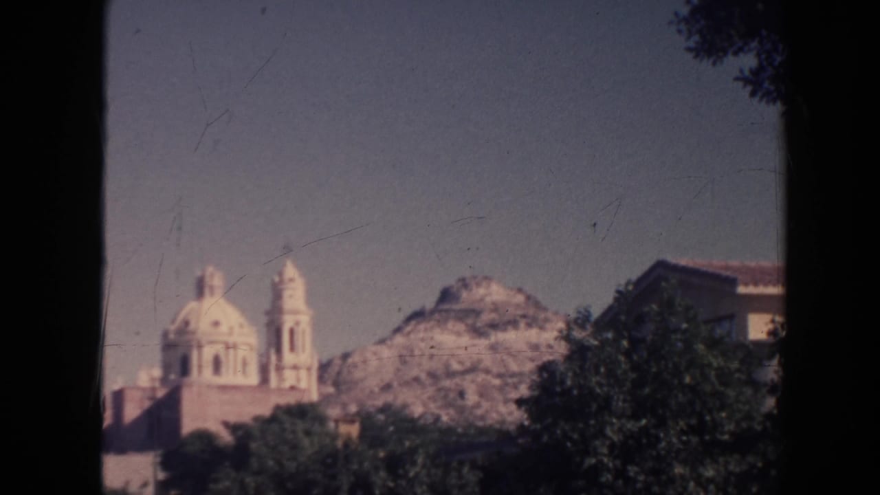 Distant pale church with domes and towers behind trees, with a rocky hillside beyond.