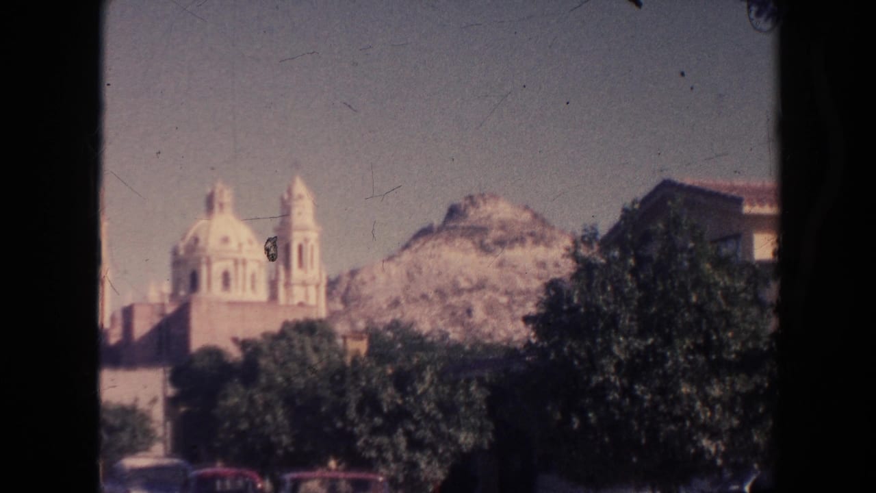 Church-like building with domes, trees in front, hillside behind, and parked cars at the bottom edge.