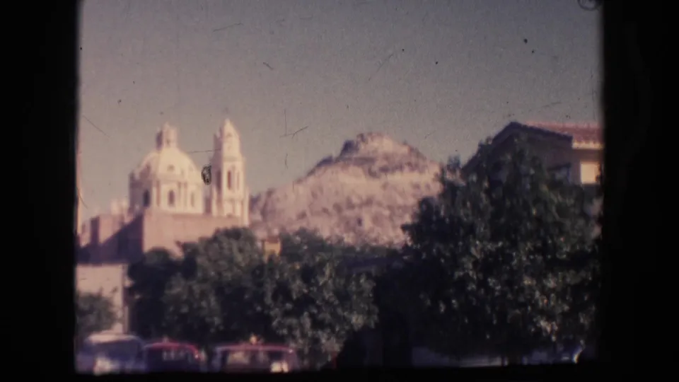 Faded 1961 8mm frame of a distant church-like landmark in Hawaii, with trees, parked cars, hillside, and visible film wear.