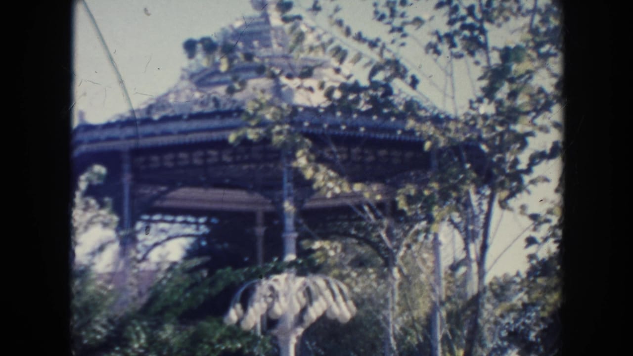 1961 Hawaii home-movie frame showing a domed gazebo behind leafy trees and shrubs in a sunlit park.