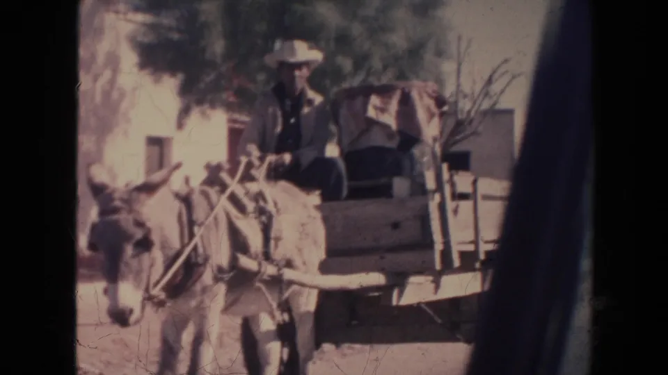 1960s home-movie frame of a man in a white hat guiding a donkey-drawn cart past buildings and trees.