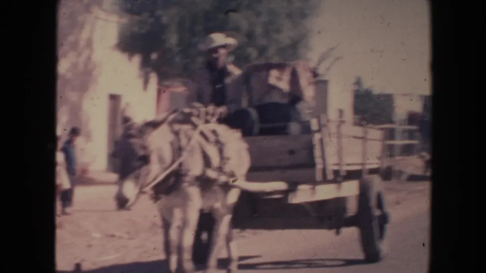 1960s 8mm frame with a donkey cart, rider, cart load, simple building, and a distant pedestrian on the street.