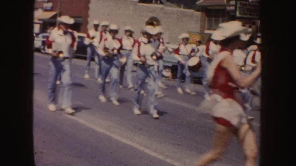 Band members in white shirts, red vests, jeans, and cowboy hats march along a South Dakota street during the supplied 1954 parade scene.