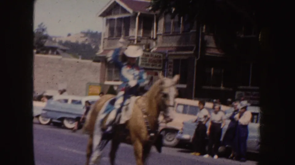 A rider in a blue-and-white costume rides a palomino horse past spectators, parked mid-century cars, and storefronts in the 1954 South Dakota parade footage.