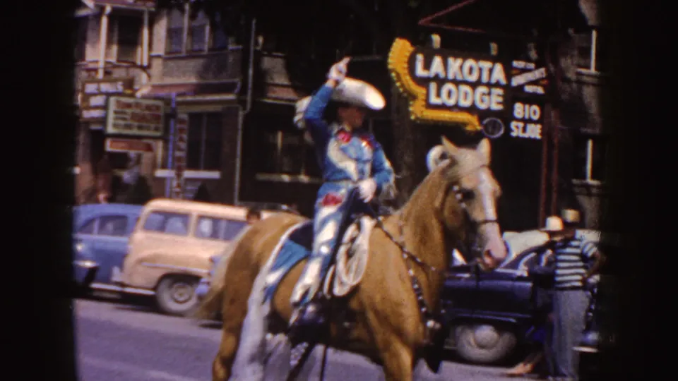 A blue-and-white costumed rider on a palomino horse passes a visible Lakota Lodge sign, parked cars, and parade spectators in South Dakota footage from 1954.