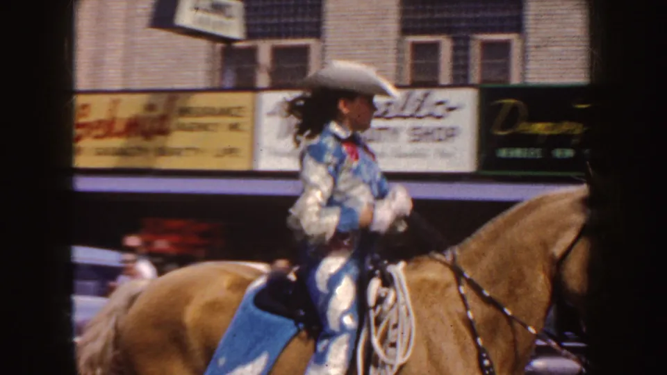 A woman in Western parade dress rides a palomino horse along a South Dakota street, with signs and cars behind her.