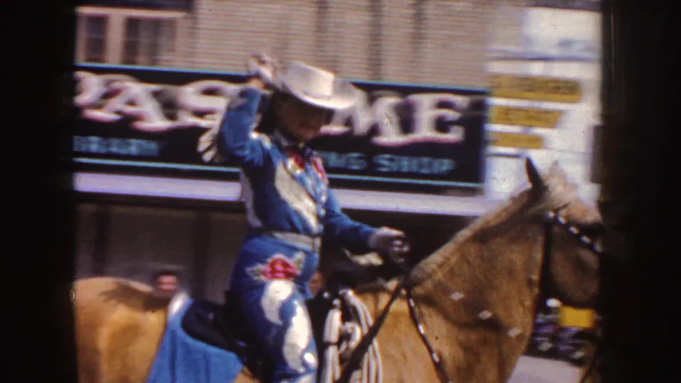 Close parade view of the woman on horseback in a sequined rodeo outfit, with a storefront sign and street activity behind her.