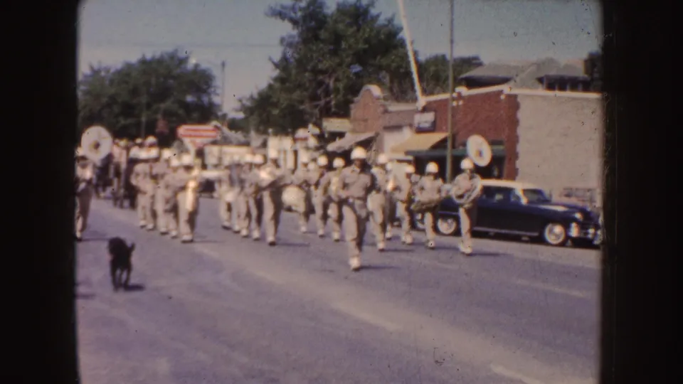 A marching band in uniform moves along a South Dakota town street during the same 1954 parade sequence.
