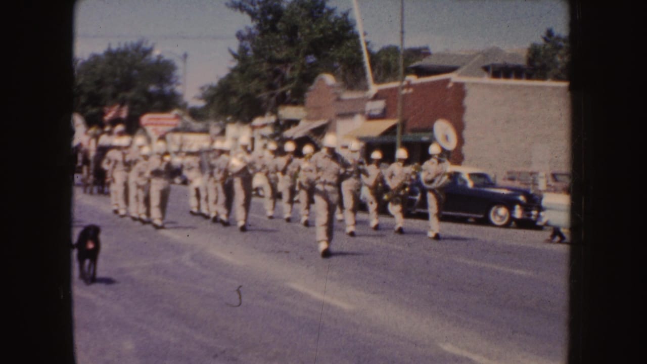 Marching band in khaki uniforms moves down a town street with a black dog nearby.