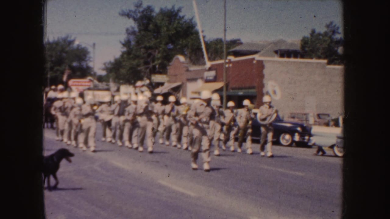 1954 South Dakota parade frame with khaki-uniformed musicians marching in formation, a black dog in the roadway, spectators, and mid-century cars nearby.