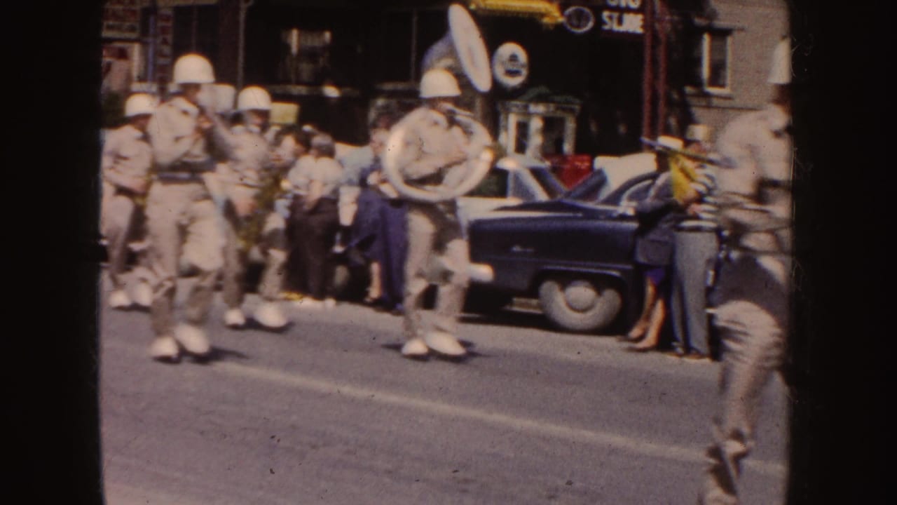 Street-level 1954 South Dakota home-movie view of marching musicians carrying a large drum and brass instruments past spectators and a dark parked car.