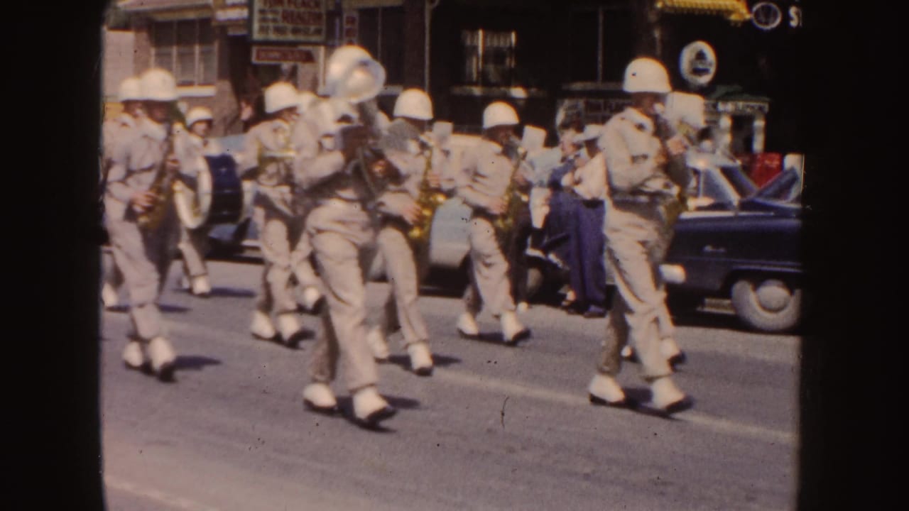 1954 South Dakota parade frame showing khaki-uniformed band members with brass instruments and drums moving past spectators, storefronts, and a parked car.