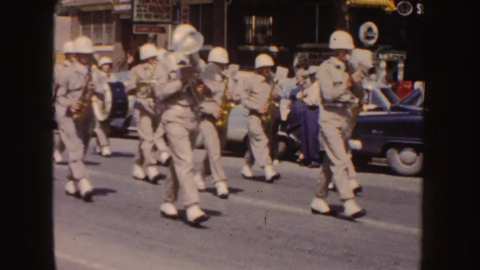 1954 South Dakota parade frame showing khaki-uniformed band members with brass instruments and drums moving past spectators, storefronts, and a parked car.