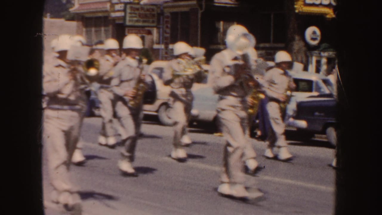 Uniformed marching band members play brass and saxophones on a sunny street.