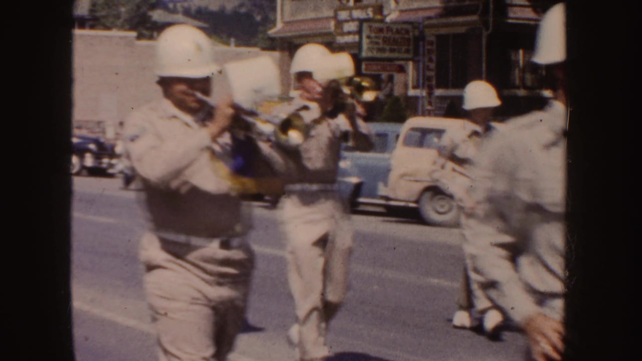 Trumpet players in light uniforms march past mid-century cars and visible storefront signs in South Dakota parade footage.