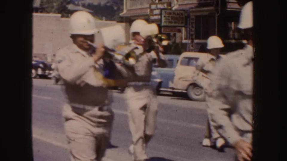Trumpet players in light uniforms march past mid-century cars and visible storefront signs in South Dakota parade footage.