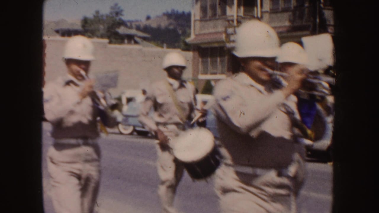 Marching band members in light uniforms pass along a town street.