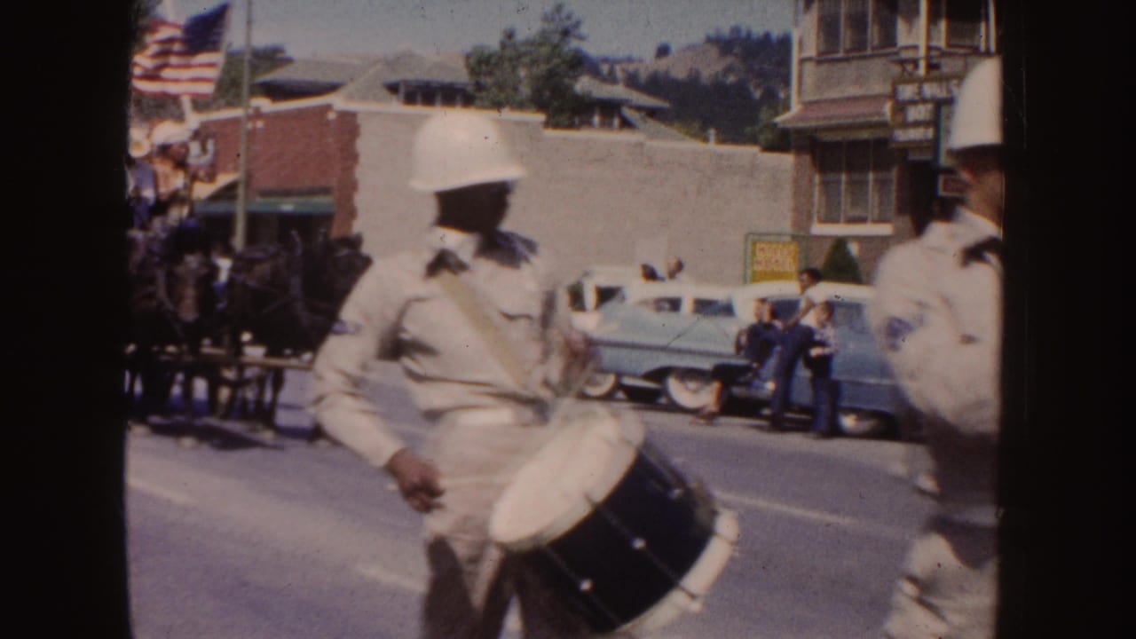1954 South Dakota parade frame with a drummer in a white helmet, an American flag, spectators, storefronts, and mid-century cars in the background.